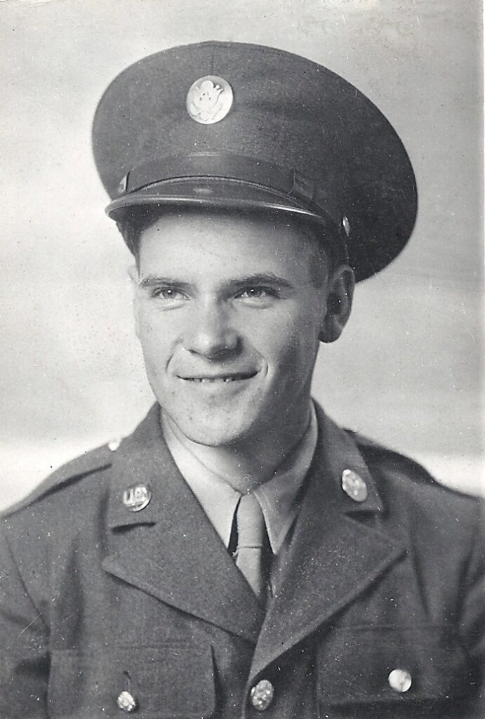 Black and white portrait, head and shoulders of a young man in a military uniform with a military hat that has a shiny button on the center of it. He is smiling.